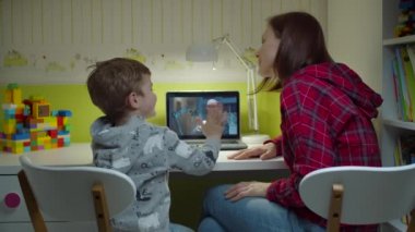 Genç bir anne, anaokulu çocuğuna dizüstü bilgisayarındaki erkek öğretmenle ödev yapmasına yardım ediyor. Evde internet eğitimi. Ebeveynler ve çocuklar birlikte okuyorlar.. 