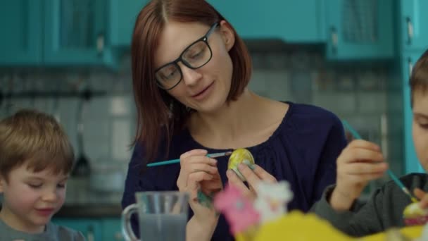 Молодая 30-летняя мать с двумя сыновьями рисует пасхальные яйца дома на голубой кухне. Счастливая семья готовится к празднованию Пасхи с цветными яйцами и шоколадным кроликом. Закрыть — стоковое видео