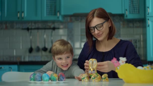 Moeder uit de 30 met zoon die thuis chocoladekonijntjes eet op de blauwe keuken. Gelukkige familie viert Pasen met gekleurde eieren en chocolade konijnen. — Stockvideo