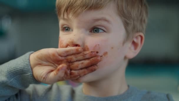 Niño rubio comiendo chocolate con la cara sucia y las manos en casa en la cocina azul. Retrato de niño feliz disfrutando de chocolate con leche en cámara lenta . — Vídeos de Stock
