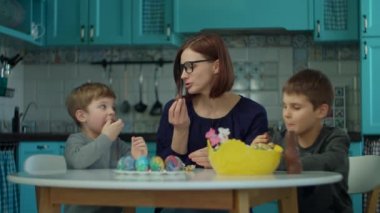 30 'ların annesi ve iki oğlu mavi mutfakta çikolatalı tavşan yiyor. Mutlu aile Paskalyayı renkli yumurtalar ve çikolatalı tavşanlarla kutluyor.. 