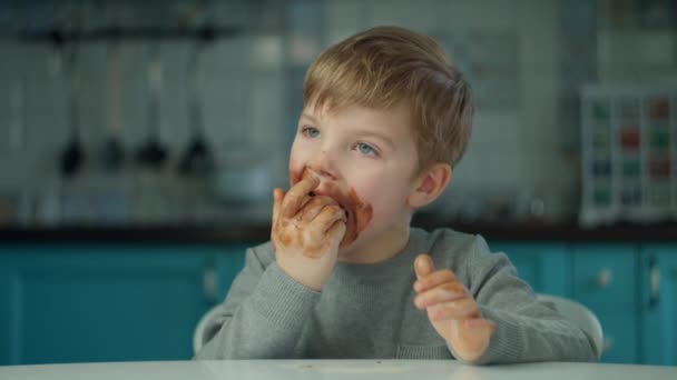 Blondine jongen eet paashaas met vies gezicht thuis op de blauwe keuken. Gelukkig kind viert Pasen met gekleurde eieren en chocolade konijntjes. — Stockvideo