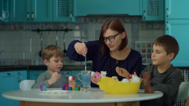 Jeune mère des années 30 avec deux fils peignant des œufs de Pâques à la maison sur cuisine bleue. Bonne famille se préparant pour la célébration de Pâques avec des œufs colorés et lapin en chocolat . — Video