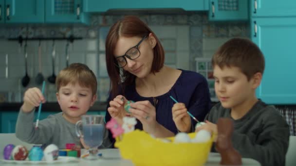 Joven madre de 30 años con dos hijos pintando huevos de Pascua en casa en la cocina azul. Familia feliz preparándose para la celebración de Pascua con huevos de colores y conejito de chocolate . — Vídeos de Stock