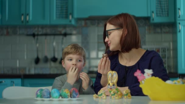30-letnia matka z synem jedzącym czekoladowe króliczki w domu w niebieskiej kuchni. Szczęśliwa rodzina świętuje Wielkanoc z kolorowymi jajkami i czekoladowymi króliczkami. — Wideo stockowe