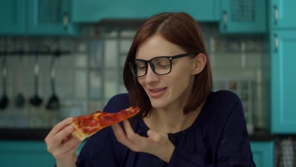Eine junge Frau in den 30er Jahren isst zu Hause Pizza in der blauen Küche. Nahaufnahme einer Frau in Brille, die Pizza genießt und in Zeitlupe in die Kamera schaut. — Stockvideo