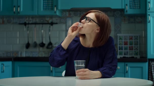 Jonge vrouw in de 30 in glazen nemen vitaminen en pillen met glas water op de keuken thuis in slow motion. — Stockvideo