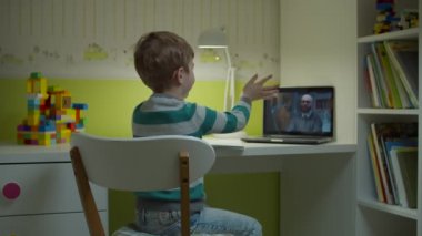 Anaokulu çocuğu dizüstü bilgisayar ekranında erkek öğretmenle çevrimiçi okul ödevi yapıyor. Çocuk evdeki çocuk odasında masada oturuyor ve internetten ders çalışıyor.. 