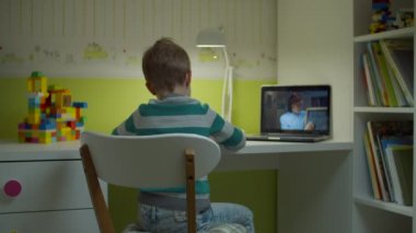 Anaokulu çocuğu dizüstü bilgisayar ekranında genç bayan öğretmenle online okul ödevi yapıyor. Çocuk evdeki çocuk odasında masada oturuyor ve internetten ders çalışıyor.. 