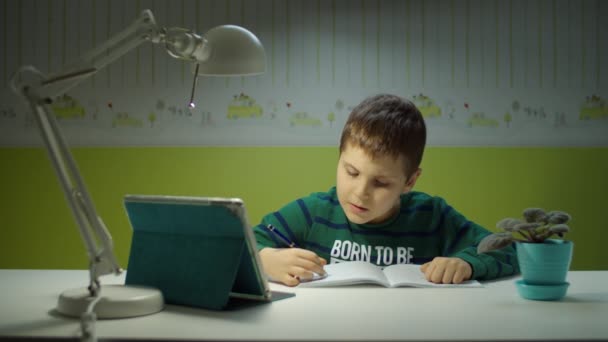 Schooler menino fazendo lição de casa on-line com computador tablet. Criança sentada na mesa e estudando on-line na sala de crianças em casa. Menino caligrafia no livro didático — Vídeo de Stock