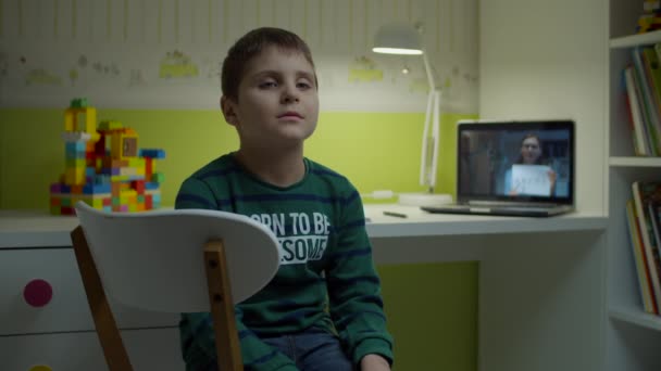 Garçon écolier ennuyé assis au bureau tandis que la leçon en ligne avec une enseignante sur l'écran de l'ordinateur portable. Éducation en ligne à la maison. Enfant autiste étudiant en ligne . — Video