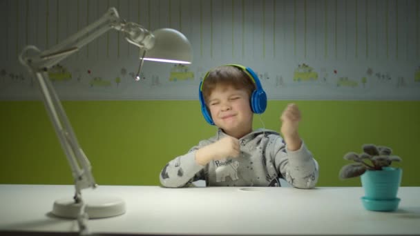 Niño preescolar en auriculares de color disfrutando de la música con los ojos cerrados sentado en el escritorio en casa . — Vídeos de Stock