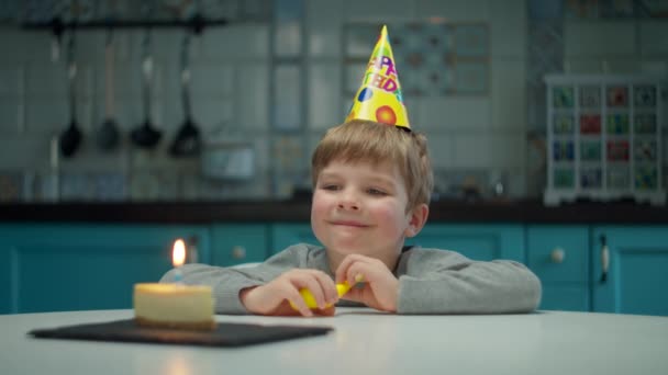 Niño preescolar joven en sombrero de cumpleaños sonriendo mirando a la cámara. Feliz cumpleaños niño solo en casa con vela en trozo de pastel . — Vídeos de Stock