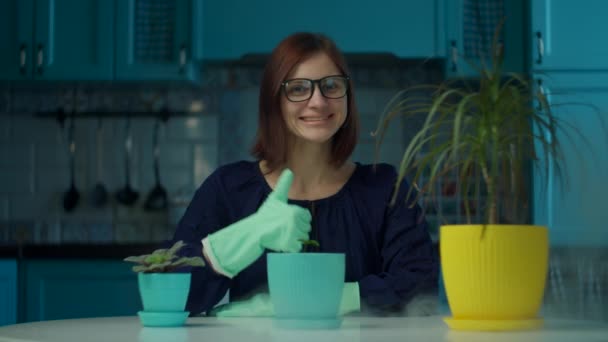 Mujer joven de 30 años en gafas y guantes haciendo pulgar hacia arriba y sonriendo. Señora disfrutando del proceso de plantación doméstica en la cocina azul — Vídeos de Stock