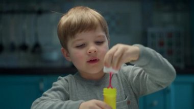 Sarışın anaokulu öğrencisi çocuk evde oturmuş sabun köpüğü üflüyor. Çocuk evde ağır çekimde tek başına oynuyor.. 