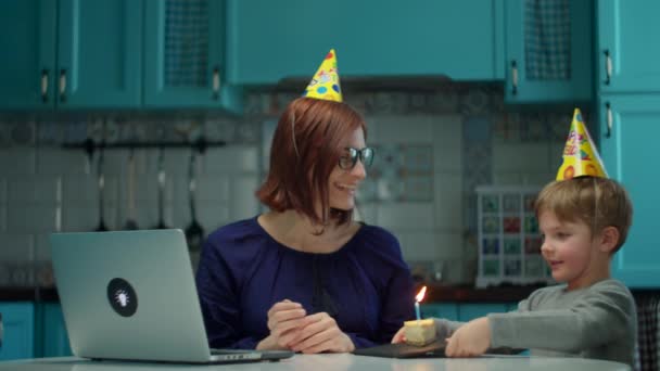 Anak laki-laki dengan topi ulang tahun membawa sepotong kue dengan lilin untuk ibu bekerja. Selamat ulang tahun wanita dengan anak di rumah di dapur. — Stok Video