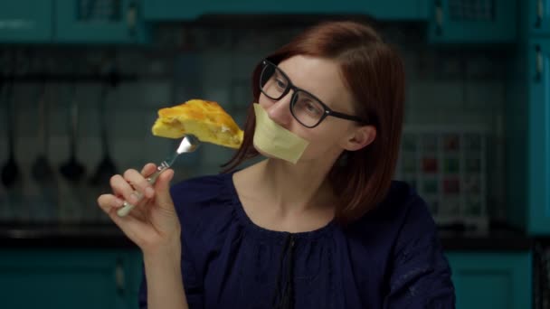 30-летняя женщина в очках с заклеенным ртом держит кусочек яблочного пирога на вилке, снимает ленту и начинает есть сладости. Лицо с ограничениями на питание . — стоковое видео