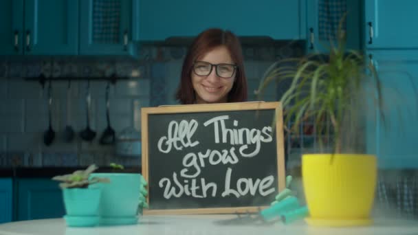 Mujer joven de 30 años con gafas y guantes sosteniendo pizarra con cantar All Things Grow With Love y sonriendo. Señora disfrutando del proceso de plantación doméstica en la cocina azul — Vídeo de stock