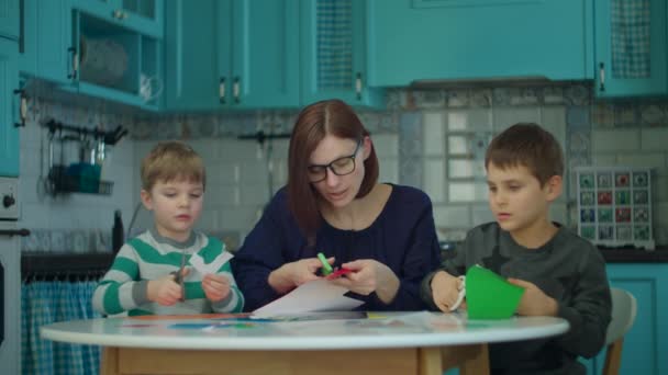 30 'lu yaşlarda, iki çocuklu genç bir anne mavi mutfakta kalem, makas ve renkli kağıtlarla yaratıcı işler yapıyor. Evde mutlu bir aile — Stok video
