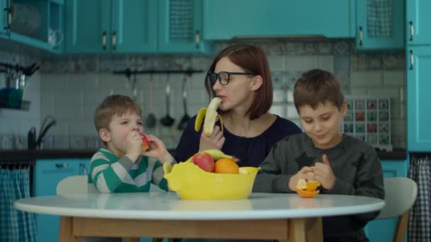 Junge 30-jährige Mutter mit zwei Kindern isst Früchte auf blauer Küche. Glückliche Familie genießt zu Hause frische Bananen, Orangen und Äpfel. — Stockvideo