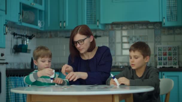 Junge Mutter in den 30er Jahren mit zwei Kindern beim kreativen Arbeiten mit Kleber, Schere und Farbpapier auf blauer Küche. Glückliche Familie zu Hause, die Kunst macht — Stockvideo