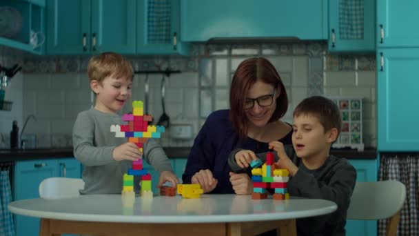 Jovem mãe dos anos 30 com dois filhos brincando com tijolos construtor na cozinha azul. Família feliz se divertir com brinquedos em casa . — Vídeo de Stock