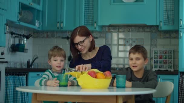 Jeune mère des années 30 avec deux enfants buvant du jus de fruits assis à la table avec des fruits sur la cuisine bleue. Bonne famille profiter de la boisson à la maison . — Video