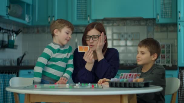 Joven madre de 30 años con dos niños jugando juegos de mesa en la cocina azul. Familia feliz divertirse con juegos de cartas en casa . — Vídeos de Stock