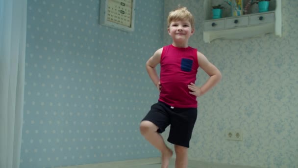 Kleuterjongen maakt thuis complex van fitnessoefeningen. Kind in rood t-shirt doet thuis sport warm up kijken naar de camera in slow motion. — Stockvideo