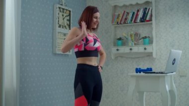 30 'lu yaşlarda spor giyimli bir kadın dizüstü bilgisayarın önünde dikilip evde monitörle konuşuyor. Kadın, evde yavaş çekimde spor dersi veriyor.. 
