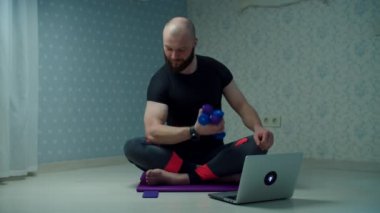 Spor giyimli 30 'lu yaşlardaki sakallı adam evde dizüstü bilgisayar kullanarak spor minderinde halterlerle spor egzersizi yapıyor. Adam ağır çekimde spor yapıyor.. 