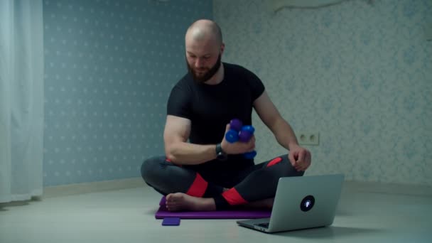 Spor giyimli 30 'lu yaşlardaki sakallı adam evde dizüstü bilgisayar kullanarak spor minderinde halterlerle spor egzersizi yapıyor. Adam ağır çekimde spor yapıyor.. — Stok video