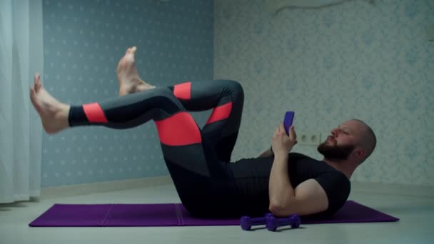 Vousatý třicátník ve sportovním oblečení, aby cyklistické sportovní cvičení na fitness rohožce pomocí mobilního telefonu doma. Muž dělá doma fitness cvičení s činky ve zpomaleném filmu. — Stock video