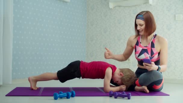 Ung mamma i sportkläder hjälper förskolebarn att träna planka hemma. Lycklig familj som tränar hemma med hantlar i slow motion. — Stockvideo