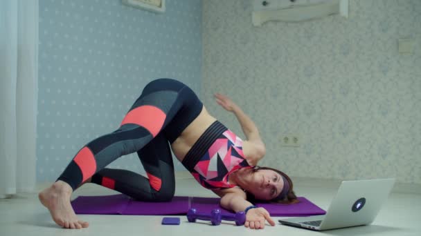 Mulher de 30 anos em sportswear fazendo ioga e exercícios de alongamento no tapete de fitness em casa. Mulher fazendo exercícios de fitness em casa usando laptop em câmera lenta . — Vídeo de Stock