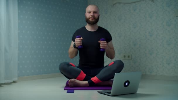 Бородатий 30-х чоловік у спортивному одязі робить вправи для фітнесу з гантелями на спортивному килимку, використовуючи ноутбук вдома. Чоловік робить домашні фітнес вправи в повільному русі . — стокове відео