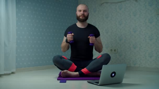 Barbudo hombre de 30 años en ropa deportiva haciendo ejercicio de fitness con mancuernas en la alfombra deportiva utilizando el ordenador portátil en casa. Hombre haciendo ejercicios de fitness en casa en cámara lenta . — Vídeos de Stock