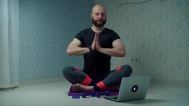 Spor giyimli 30 'lu yaşlarda yoga yapan ve spor paspası üzerinde egzersiz yapan sakallı bir adam evde dizüstü bilgisayar kullanarak spor yapıyor. Adam yavaş çekimde halterlerle spor yapıyor.. 