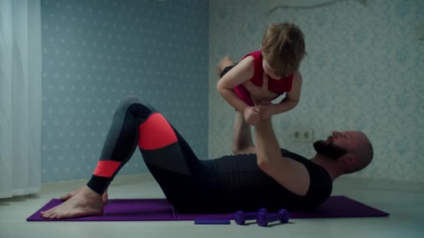 Vousatý třicátník otec ve sportovním oblečení dělat kliky cvičení s malým synem na rukou. Šťastná rodina dělá fitness společně doma. — Stock video