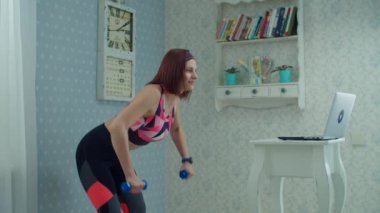 30 'lu yaşlarda kadın spor kıyafetleri içinde dizüstü bilgisayarın önünde duruyor ve ağır çekimde halterlerle kol ve kol kası egzersizleri yapıyor.. 
