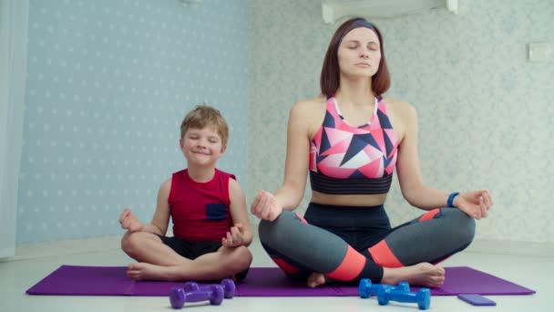 30-letnia matka w odzieży sportowej ćwiczy medytację z młodym chłopcem na macie fitness w domu. Szczęśliwa rodzina medytuje razem w zwolnionym tempie. — Wideo stockowe