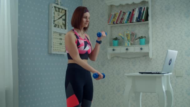 30s femme en vêtements de sport debout devant un ordinateur portable et faire des exercices biceps et triceps avec haltères à la maison au ralenti . — Video