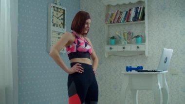 30 'lu yaşlardaki spor elbiseli kadın dizüstü bilgisayarın önünde duruyor ve evde spor egzersizleri yapıyor. Dambıllarla spor yapan kadın yavaş çekimde evde.. 
