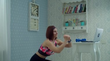 30 'lu yaşlarda kadın spor kıyafetleri içinde dizüstü bilgisayarın önünde duruyor ve evde çömelme egzersizleri yapıyor. Kadın evde ağır çekimde spor yapıyor.. 
