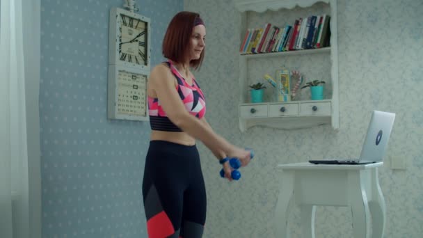 30s femme en vêtements de sport debout devant un ordinateur portable et faire des exercices biceps et triceps avec haltères à la maison au ralenti . — Video