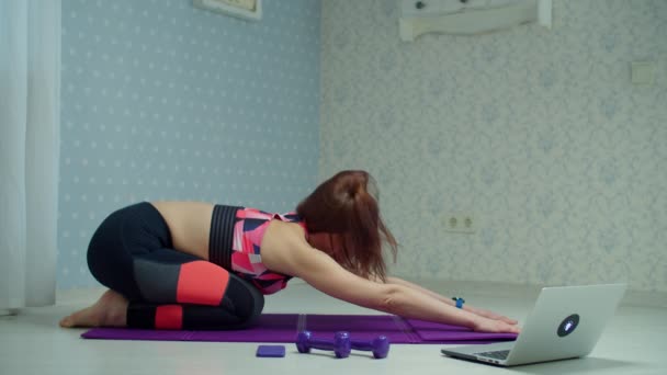 Mulher de 30 anos em sportswear fazendo ioga e exercícios de alongamento no tapete de fitness em casa. Mulher fazendo exercícios de fitness em casa usando laptop em câmera lenta . — Vídeo de Stock