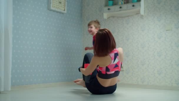 30-летняя мать в спортивной одежде играет с маленьким мальчиком на полу дома. Счастливая семья делает фитнес вместе в замедленной съемке . — стоковое видео