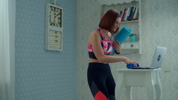 30er-Jahre-Frau in Sportbekleidung mit Fitnessmatte und Hanteln in den Händen, die zum Laptop kommen und zu Hause in Zeitlupe Fitnessübungen starten. — Stockvideo