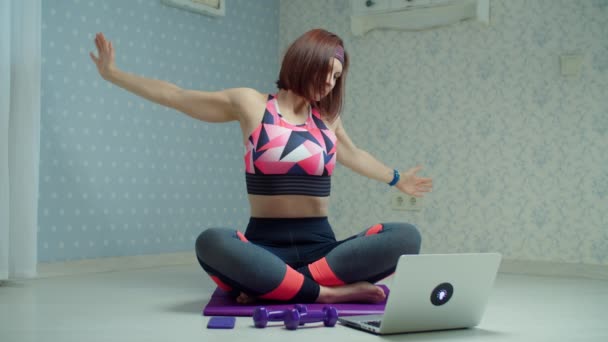 Молодая женщина 30-х годов в спортивной одежде делает йогу и упражнения на растяжку на фитнес коврик дома. Женщина делает домашние упражнения с помощью ноутбука в замедленной съемке . — стоковое видео