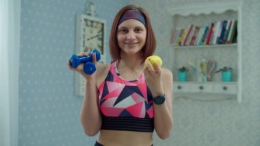 Spor giyimli 30 'lu yaşlarda, elinde dambıllar ve elinde elma meyveleri olan genç bir kadın yavaş çekimde kameraya bakıyor. Spor, yoga ve spor ev konsepti. 